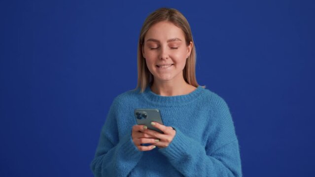 Positive woman wearing blue sweater texting by phone in blue studio