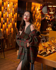 Fototapeta premium Joyful young woman with red lipstick and loose flowing brown hair in green checkered coat holds glass of wine dancing in cozy pub