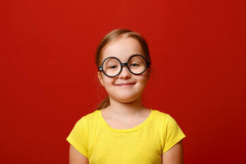 Funny little girl with glasses. Schoolgirl child in a yellow t-shirt on a red isolated background.