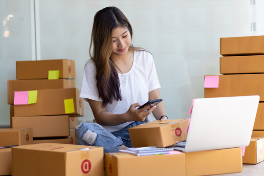 A woman running a small online business holds a mobile phone taking pictures of parcels sent to customers.