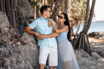 Obraz na płótnie Canvas Adorable pretty man and a woman in sunglasses and ordinary clothes on the background of the sea posing near a stone under a palm tree and looking at each other. Romance concept.