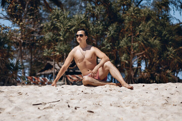 Lovely handsome young man in beach clothes and sunglasses posing sitting on the sand against the background of palm trees. Vacation concept.