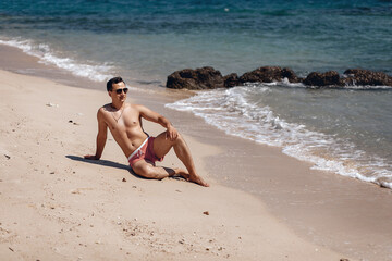 Rich amazing sexual guy in swimming clothes lying on the beach next to the water, stones and sand. Concept of relaxion.