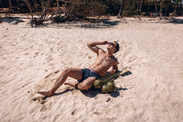 Attractive satisfied man lying on the sand, smiling and sunbathing next to coconuts. Concept of relaxion