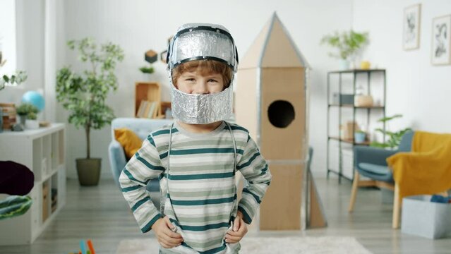 Portrait of cheerful little child wearing astronaut cosume smiling in room with handmade cardboard spaceship enjoying game at home. Childhood and leisure concept.