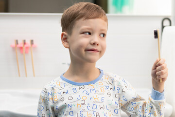 A cute seven-year-old boy in pajamas with a bamboo toothbrush in his hand before going to bed at home in the bathroom against the backdrop of a washbasin with a faucet. Selective focus. Portrait