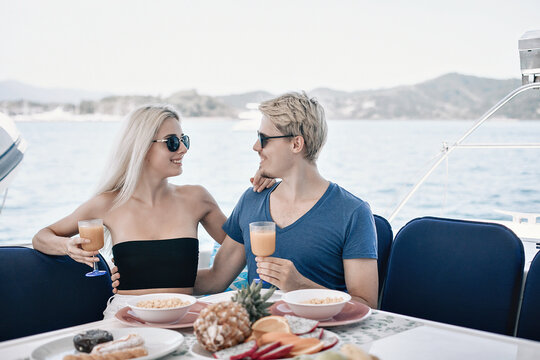 Lovely young and pretty couple man and woman are having dinner at the table of their huge and expensive white yacht, smiling and holding glasses of juice in their hands, enjoying each other's company