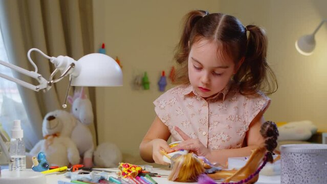 Cute little girl is sewing a button to the doll's clothes. Child learns sewing skills and clothing design while sitting at the table in the bedroom.