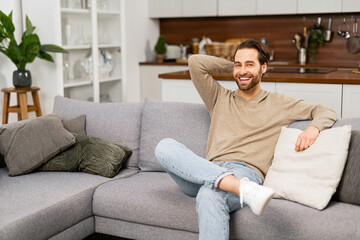 Portrait of young caucasian man resting on sofa, thinking about the future. Handsome positive man with hand on the head sitting in living room and relaxing at home