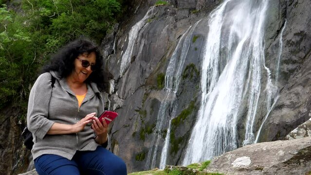 Adult woman sitting on the rock by waterfall enjoying fresh pictures tilt up shot.