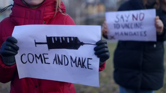 Two female protestors with Come and make banner and Say no to vaccine mandates banners outdoors. Unrecognizable Caucasian women on coronavirus pandemic rioting on autumn spring day