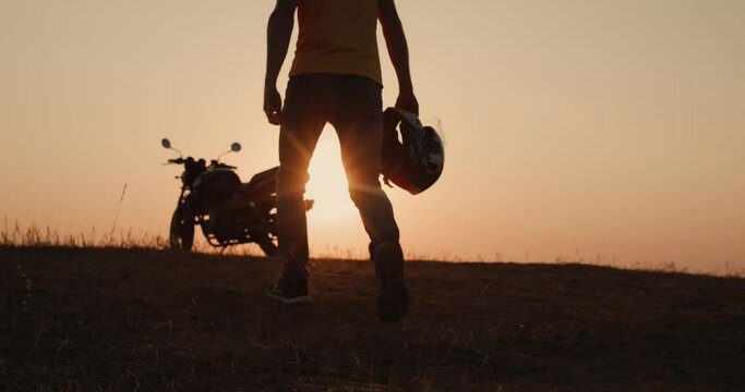 Start the journey - a man with a helmet in his hand goes to the motorcycle at sunset