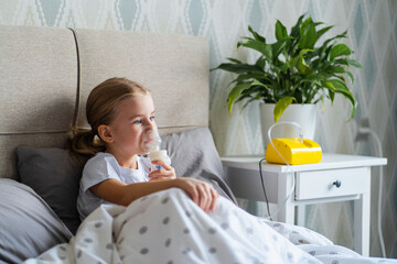 Sick little girl making inhalation with nebulizer to reduce coughing, lying in bed at home, child...