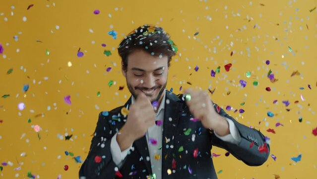 Portrait of Middle Eastern dancer moving while confetti falling down in air during holiday celebration. Man in elegant suit dancing on yellow background.