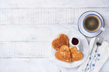Pancakes with berry jam and honey in shape of heart and hot cup of coffee over white wooden background. Concept  breakfast for Valentines Day or pleasant surprise for loved one Table viewed from above