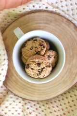 Mug filled with chocolate chip cookies and soft knitted blanket on a bed. Flat lay.
