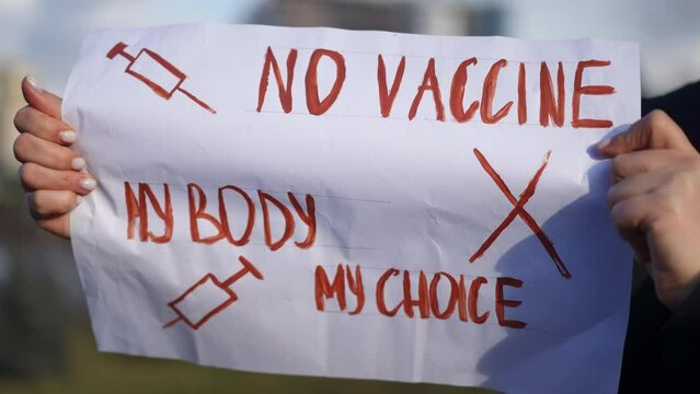 Close-up anti-vax placard in Caucasian female hands with blurred urban city at background. Unrecognizable woman protesting against mandatory Covid-19 vaccination and green pass outdoors