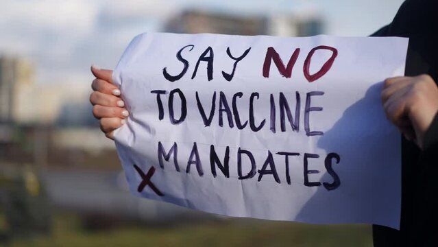 Say no to vaccine mandates placard in female Caucasian hands with blurred urban city at background. Unrecognizable serious confident protestor against mandatory coronavirus vaccination outdoors