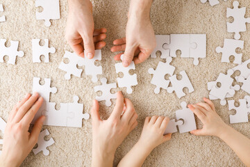 Mother, father and toddler hands playing and assembling white puzzle pieces on light beige carpet...