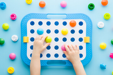 Baby boy hands playing and putting colorful plastic buttons in holes of white board. Blue table background. Pastel color. Closeup. Children development with color cognition game. Top down view.