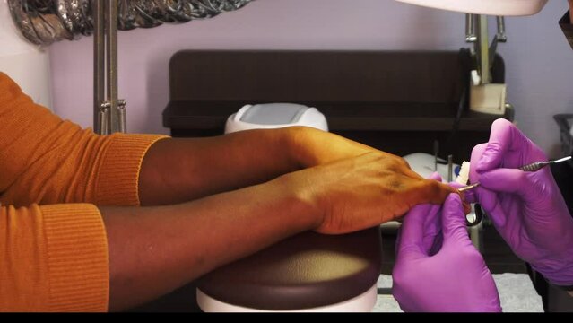 Female master in protective gloves makes manicure to black man in a salon in closeup. She removes cuticle by a metal pusher. African american person takes care of his hands coming to beauty procedures