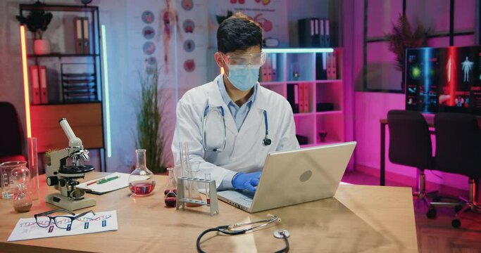 Attractive concentrated hardworking qualified man scientist in protective mask working on laptop during overtime work in evening laboratory