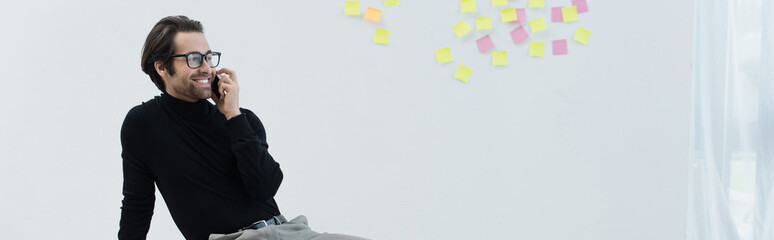 pleased man in eyeglasses talking on mobile phone near sticky notes on grey wall, banner.