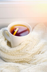 Fototapeta na wymiar A cup of hot tea with lemon is wrapped in a white knitted scarf. Warm cozy atmosphere. Soft focus, vertical photography.