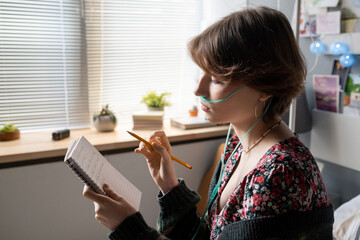 Teenage girl with pencil looking through her notes in notebook while sitting on bed in front of...