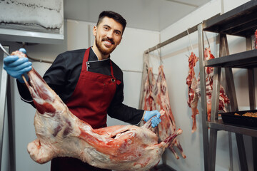 Male butcher standing by meat hanging on hook in meat shop