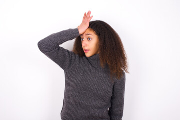 teenager girl wearing grey sweater standing against wite background surprised with hand on head for...