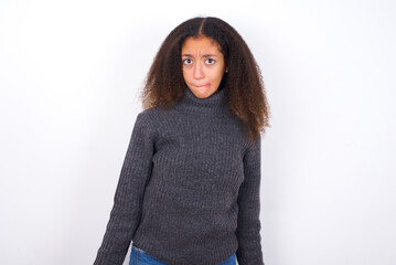 teenager girl wearing grey sweater standing against wite background making grimace and crazy face,...