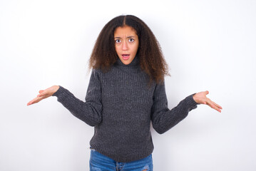 Frustrated teenager girl wearing grey sweater standing against wite background feels puzzled and hesitant, shrugs shoulders in bewilderment, keeps mouth widely opened, doesn't know what to do.