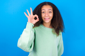 Fototapeta premium beautiful teenager girl wearing green sweater standing against blue background hold hand arm okey symbol toothy approve advising novelty news