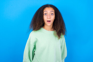 Fototapeta premium Shocked beautiful teenager girl wearing green sweater standing against blue background stares bugged eyes keeps mouth opened has surprised expression. Omg concept