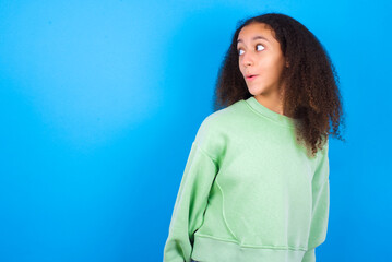Fototapeta premium Shocked beautiful teenager girl wearing green sweater standing against blue background look empty space with open mouth screaming: Oh My God! I can't believe this.