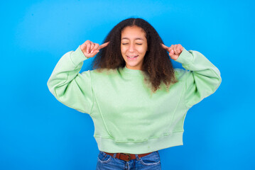 Photo of crazy beautiful teenager girl wearing green sweater standing against blue background screaming and pointing with fingers at hair closed eyes