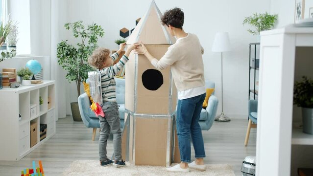 Happy young family mother and son making cardboard rocket playing together in cozy apartment. Futuristic toy and inspiration concept.