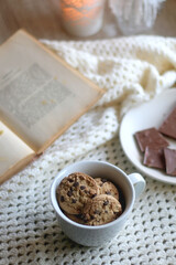 Plate with almond chocolate bar, mug with chocolate chip cookies, open book, reading glasses, soft blanket and lit candles. Hygge at home. Selective focus.
