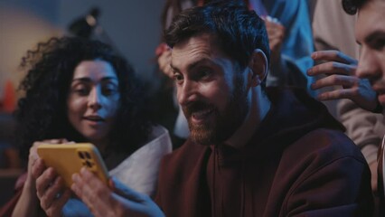 Agitated guy rejoicing while watching over sports betting at the smartphone screen. Focus on the man with device smiling and looking at the friends while sitting on sofa. Sport fans concept. - Powered by Adobe