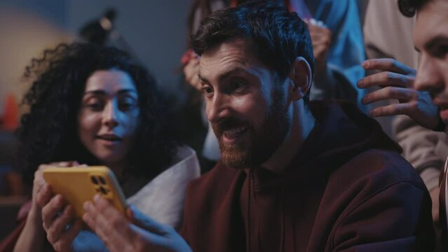 Agitated guy rejoicing while watching over sports betting at the smartphone screen. Focus on the man with device smiling and looking at the friends while sitting on sofa. Sport fans concept.
