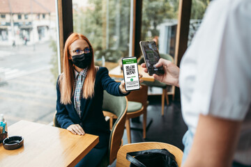 Young attractive business woman sitting in a restaurant or cafe bar and showing digital Covid-19 immunization pass certificate to the waitress. Pandemic and global security measures concept.