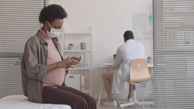 Medium stab slowmo shot of African-American pregnant woman in face mask looking at ultrasound image of her baby while sitting on medical couch in her gynecologist office