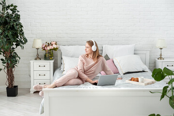 Obraz na płótnie Canvas Pleasant middle-aged blonde woman in a home suit and headphones sitting in bed with a laptop. Work and study from home. Online communication. Distance education. Space for text.