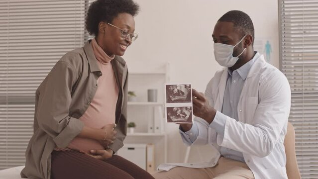 Medium slowmo shot of African-American male doctor and happy young pregnant African-American woman looking at ultrasound image of baby together during appointment