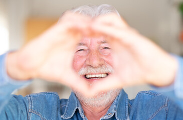 Attractive mature smiling senior man feels happy and romantic shapes heart gesture expresses tender feelings. Elderly bearded caucasian grandfather. Positive think affection and care concept