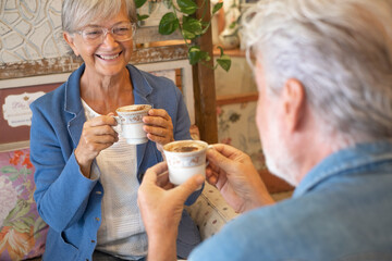 Portrait of happy adult senior couple inside coffee shop having fun and laughing drinking cappuccino. Smiling elderly people enjoying retirement and stay together, looking each other in the eyes