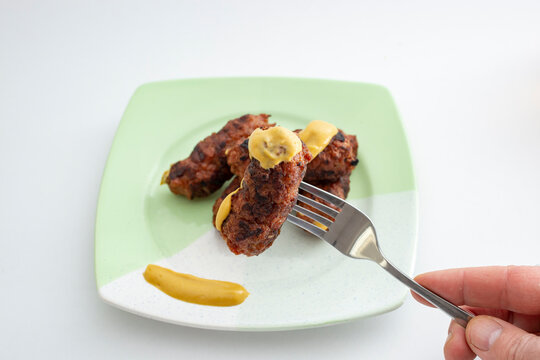 Mici or mitititei (traditional romanian food) with mustard on a ceramic plate, on white background. Hand holding a stainless steel fork