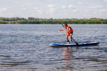 Little female child standing on paddle board on clue lake trying to keep balance on feet wearing orange life jacket. Active holidays full of adventures. Inculcation of love for sports from childhood.