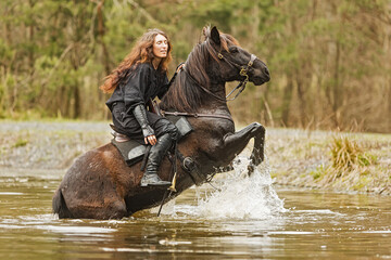 Fototapeta na wymiar black dressed young woman sitting on a horse that's on its hind legs in the lake